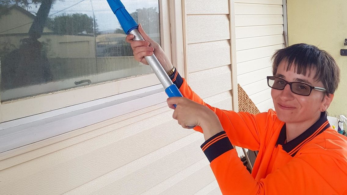 Clarissa has short brown hair, wears dark tinted glasses and an orange high vis vest. She's outside, standing near a window looking at camera, holding up a window cleaning squeagie to the window.