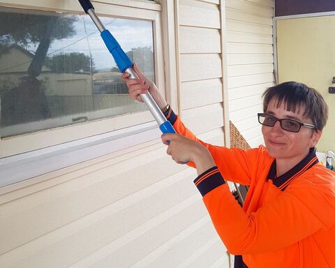 Clarissa has short brown hair, wears dark tinted glasses and an orange high vis vest. She's outside, standing near a window looking at camera, holding up a window cleaning squeagie to the window.