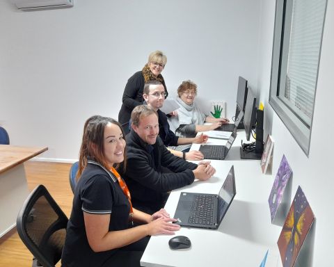 Five people in the image, three are sitting at a long desk in front of computers, and two people are poised to assist them with their work.