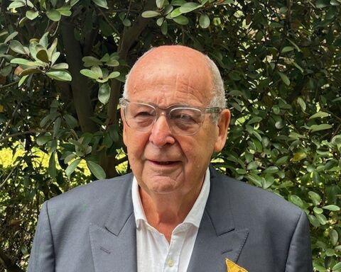 Man wearing grey suit and glasses, standing behind greenery