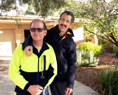 Two men, one wearing hi-vis jacket and tinted sunglasses and other wearing navy blue jacket standing next to each other putting hand on shoulder