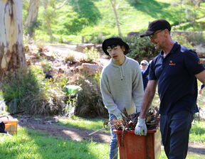 Two men wearing bucket hats holding a bucket full of branches