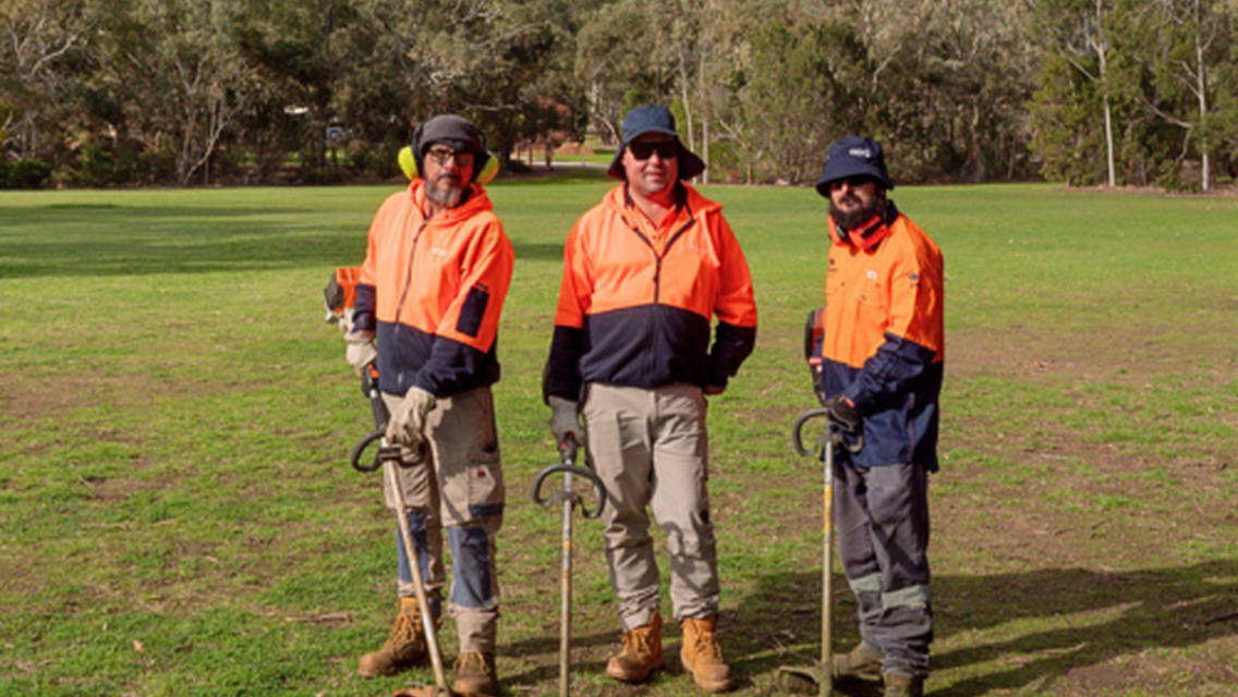 Three men wearing orange hi-vis clothing standing next to each other with gardening tools