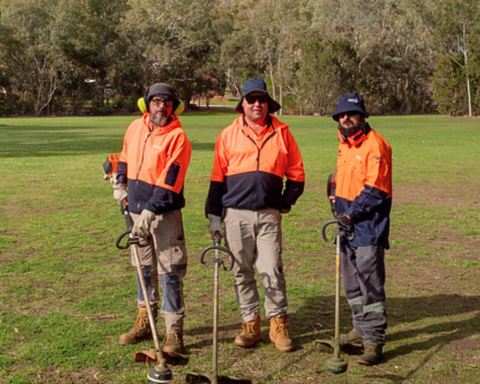 Three men wearing orange hi-vis clothing standing next to each other with gardening tools